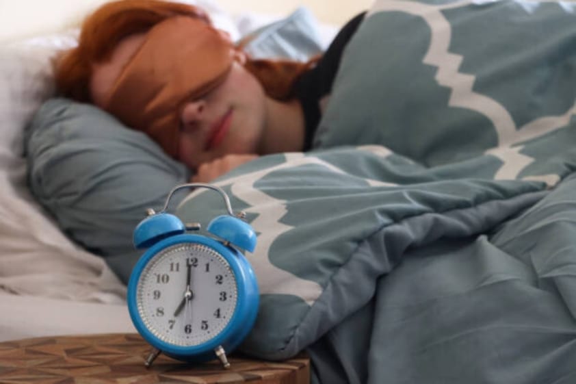 Person asleep with an eye mask and an alarm clock beside the bed.