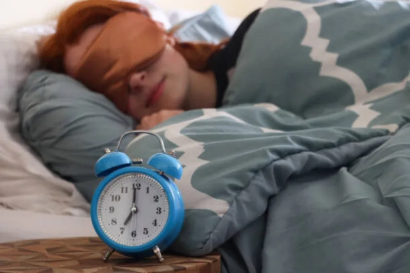 Person asleep with an eye mask and an alarm clock beside the bed.